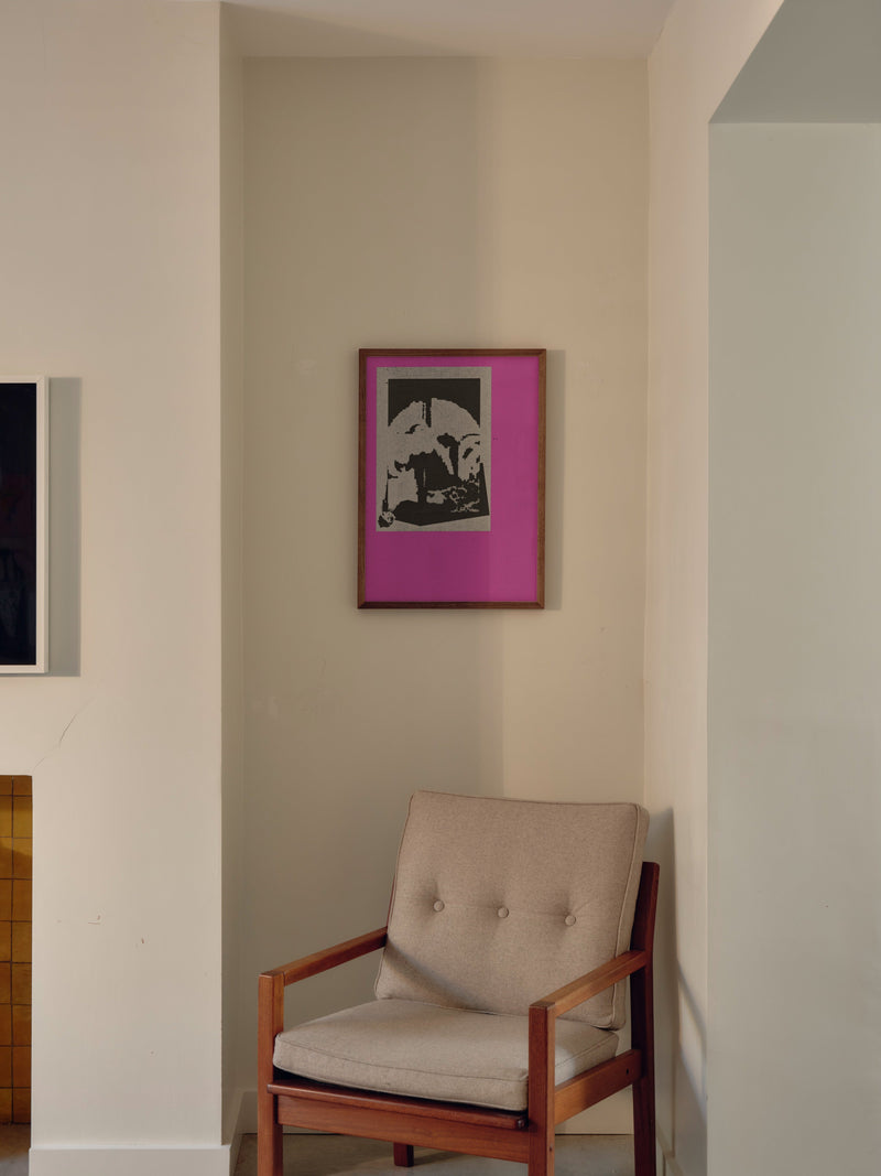 A black and white graphic print set against a pink background in a wooden frame, hung on a white wall above a small armchair. 